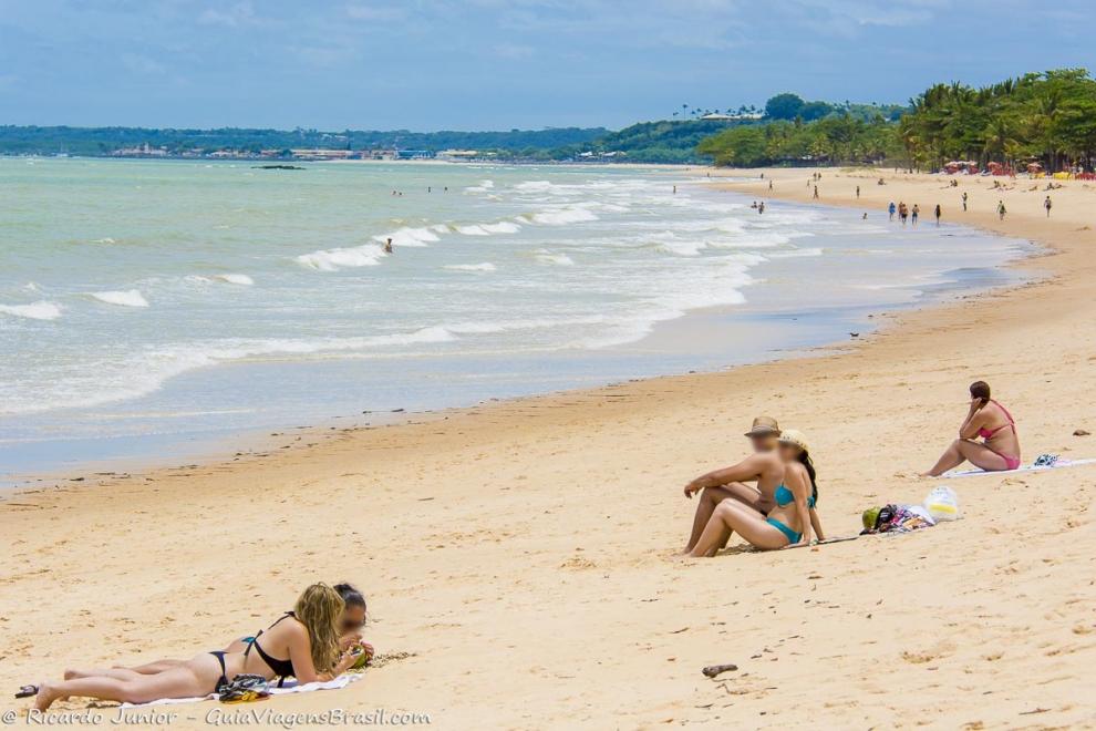 Imagem de amigas tomando sol e turistas admirando o mar.